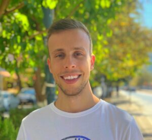 Young man smiling at camera on sunlit tree lined sidewalk with blurred cars and buildings behind him wearing a white tshirt and short light brown hair