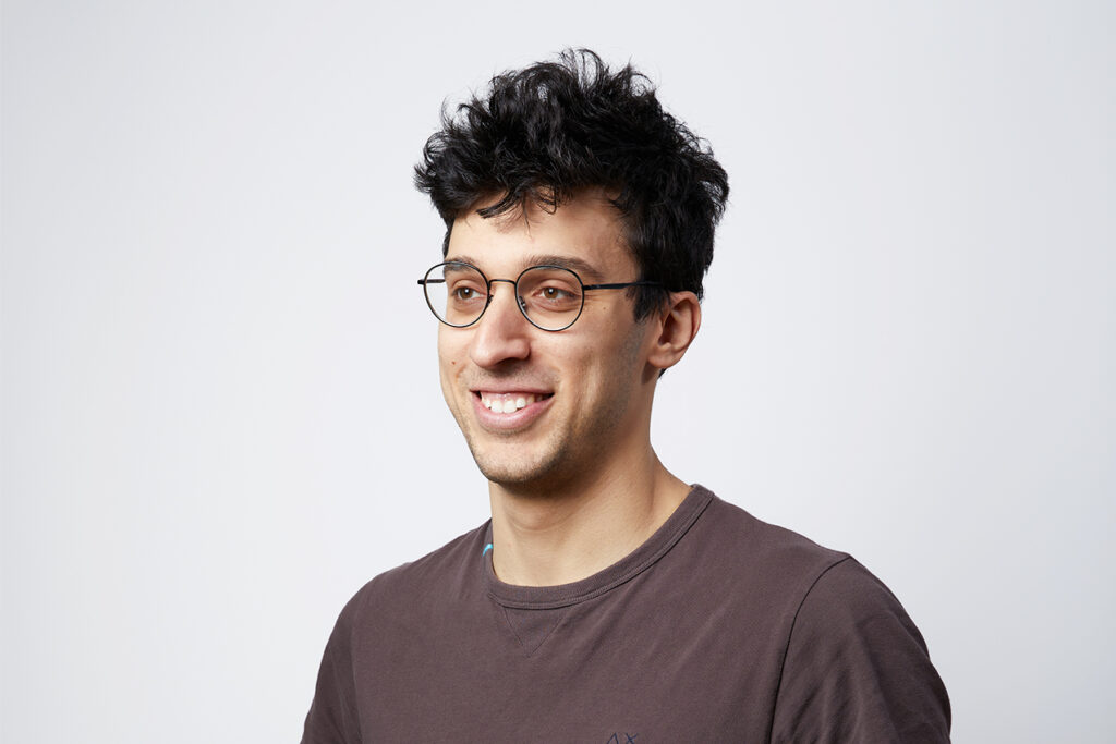 Young man wearing round glasses smiling slightly looking to the side against a plain light background with tousled dark hair and a casual brown T shirt